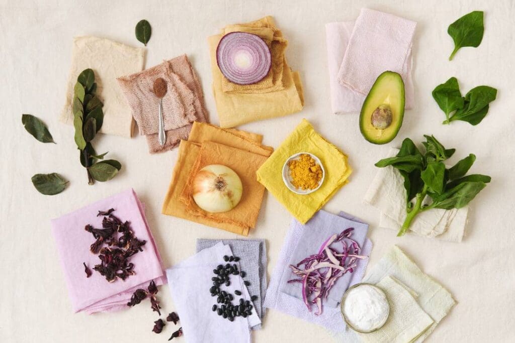 Natural ingredients like turmeric, onion skins, avocado, hibiscus, and leafy greens on colored fabrics, showcasing non-toxic, eco-friendly dyeing and sustainable kitchen practices.