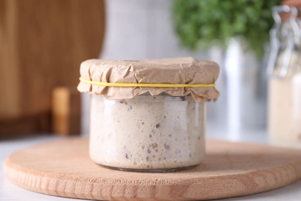 A jar of sourdough starter covered with paper and a rubber band, highlighting the natural fermentation process for making healthier bread.