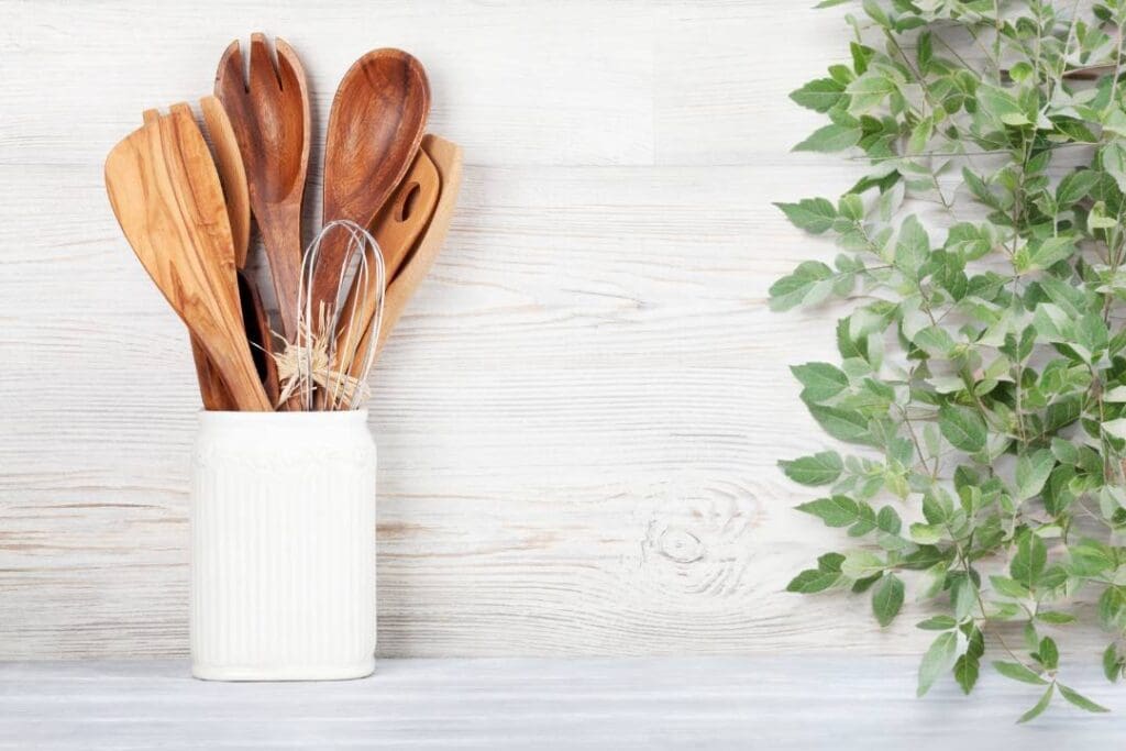 Wooden kitchen utensils in a ceramic holder, paired with greenery on a rustic white wooden background, representing sustainable and non-toxic cooking tools.