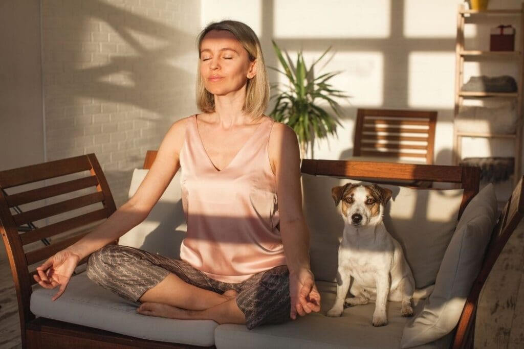 Woman meditating in the morning sunlight with her dog, practicing mindfulness and deep breathing for stress relief.