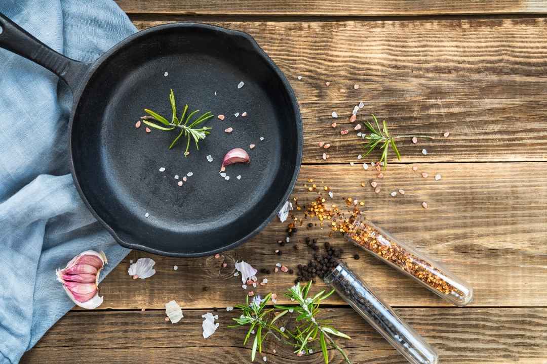 Non-toxic cookware: A cast iron skillet with fresh rosemary, garlic, and pink Himalayan salt on a rustic wooden countertop.