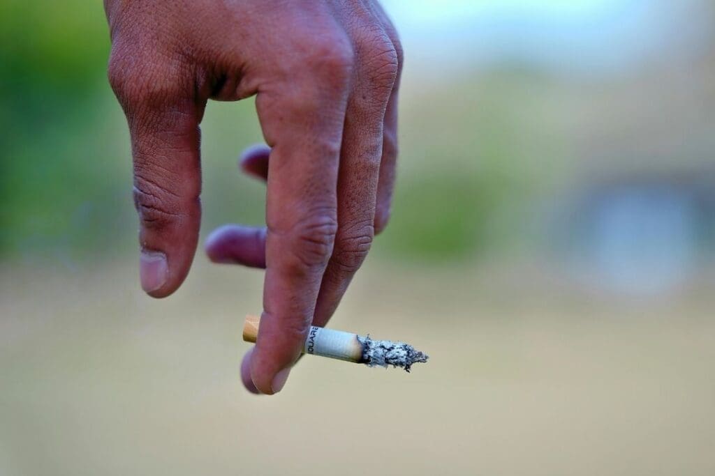 Close-up of a person's hand holding a lit cigarette, with ashes forming at the tip and smoke rising gently.